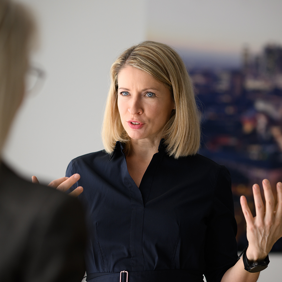 A professional woman with blond hair in a navy blue dress speaking and gesturing during a discussion.